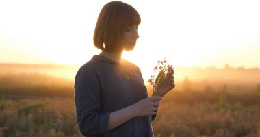 Young redhead woman in beautiful boho dress relaxing in the field during foggy sunset, female outdoors with bouquet in hands