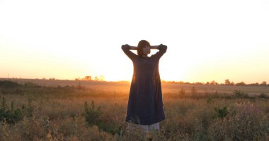 Young redhead woman in beautiful boho dress relaxing in the field during foggy sunset, female outdoors with bouquet in hands