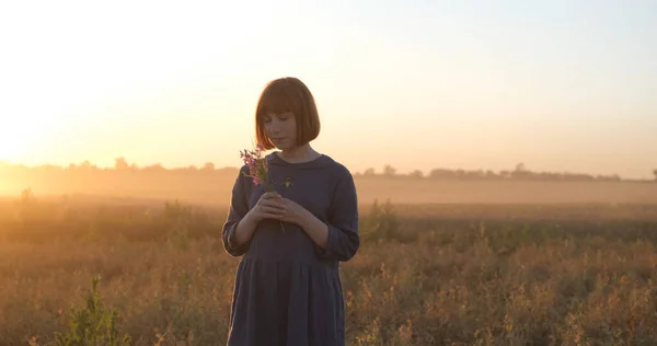 Young redhead woman in beautiful boho dress relaxing in the field during foggy sunset, female outdoors with bouquet in hands