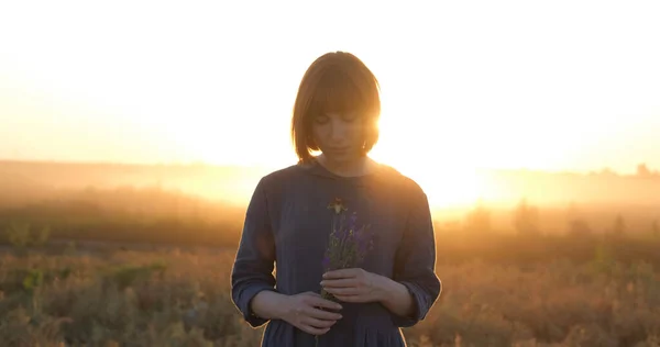 Young redhead woman in beautiful boho dress relaxing in the field during foggy sunset, female outdoors with bouquet in hands