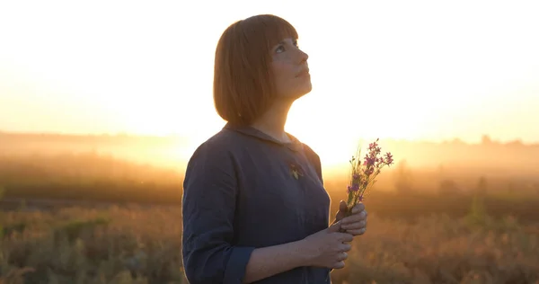 Young redhead woman in beautiful boho dress relaxing in the field during foggy sunset, female outdoors with bouquet in hands