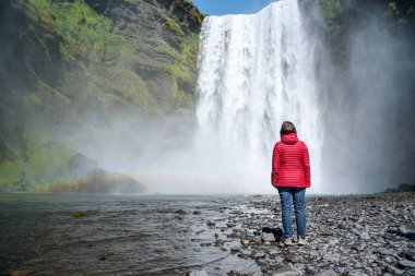 Güneşli bir yaz sabahında güçlü bir şelalenin dibinde duran kadın. Skogafoss Şelalesi, İzlanda.