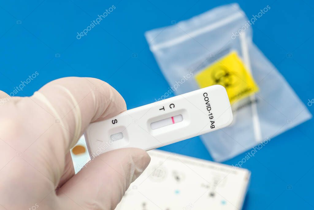 Gloved hand holding a SARS-Cov-2 antigen test strip showing a negative result. A nasal swab inside a bag is visible in background.