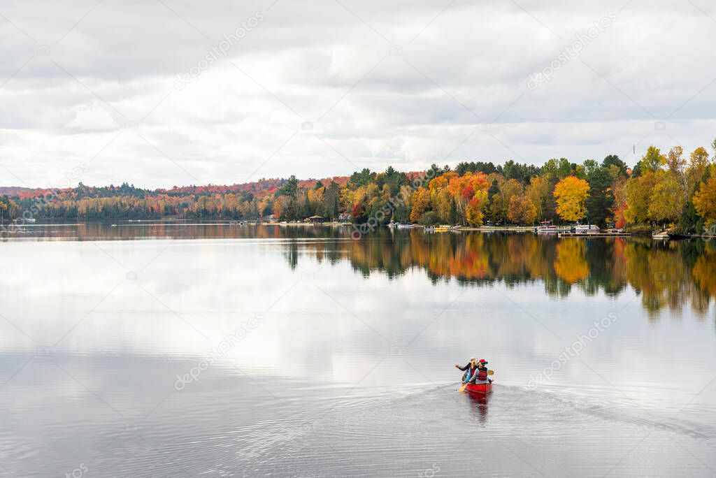 People canoeing on a beautiful lake surrounded by forest at the peak of fall foliage on a cloudy autumn day. Ontario, Canada.