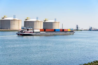 Açık bir yaz gününde büyük beton LNG tanklarının önünde konteyner mavnası satışı. Rotterdam Limanı, Hollanda.