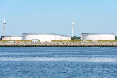 Large steel tanks in a crude oil terminal on a sunny summer day. Wind turbines are in background. Port of Rotterdam, the Netherlands.