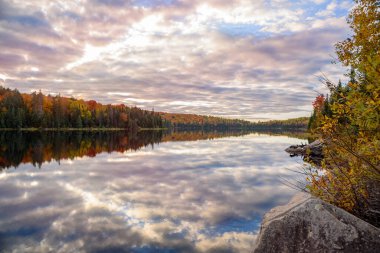 Beautiful lake with forested shores at the peak of fall foliage under cloudy sky at sunset. Reflection in water. Algonquin Park, ON, Canada.