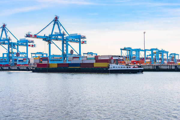 Cargo barge loaded with containers sailing past a comercial dock with tall gatry cranes and stacks of containers. Rotterdam, Nehterlands.