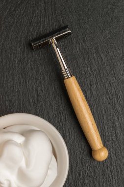 Tahta saplı bir tıraş makinesi ve taştan bir arka planda beyaz bir kasede tıraş köpüğü. Bakım için erkek aksesuarları. Düz yatıyordu.