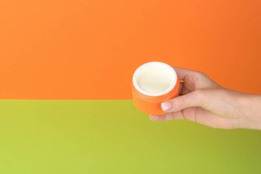 Yeşil ve turuncu arka planda krema kavanozunu tutan güzel bir kadın eli. Kozmetik kullanma kavramı.