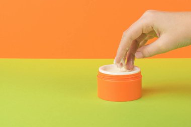 Kız yeşil-turuncu arka planda kremayla parmaklarını turuncu kavanoza daldırıyor. Kozmetik kullanma kavramı.