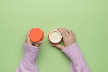 Süveter giymiş güzel bir kızın elleri yeşil arka planda bir krema kavanozunu açıyor. Kozmetik kullanma kavramı. Düz yatıyordu.