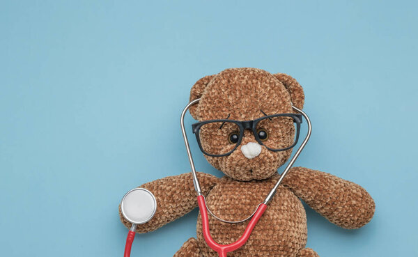 A beautiful knitted bear with a red stethoscope on a blue background. The concept of treatment of diseases.