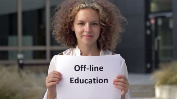 Söt tonåring flicka står med en affisch OFF-LINE UTBILDNING nära skolbyggnaden. — Stockvideo