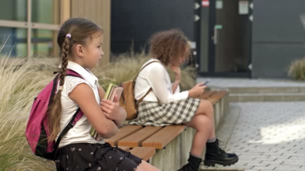 Dvě školačky různého věku sedí na lavičce na školním dvoře. Holčička si přitiskne knížky na hruď, starší má v rukou smartphone.. — Stock video