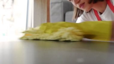 Kırmızı önlüklü ve sarı eldivenli genç bir ev hanımı rafların tozunu alıyor..