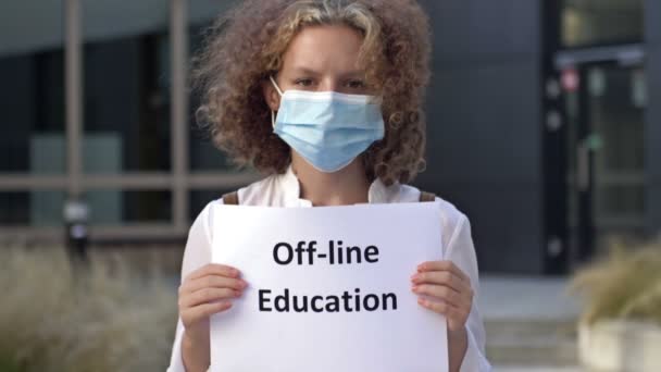Schoolgirl φορώντας μια ιατρική μάσκα κατέχει μια πινακίδα OFF-LINE ΕΚΠΑΙΔΕΥΣΗ. — Αρχείο Βίντεο
