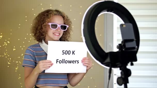Alegre blogger chica muestra un póster de 500K SEGUIDORES en la cámara. Adolescente mostrando sus logros, el crecimiento del número de suscriptores. — Vídeos de Stock