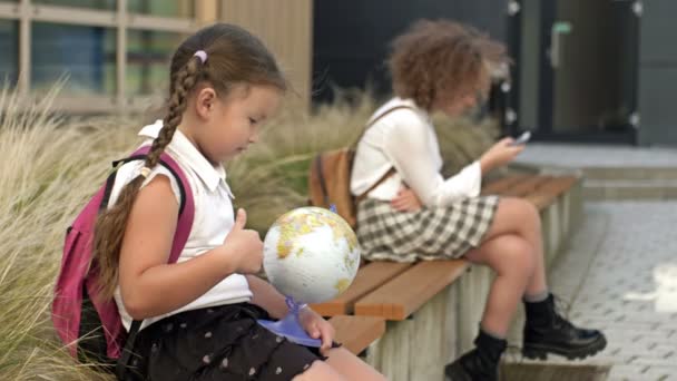 Две школьницы разного возраста сидят на скамейке во дворе школы. Маленькая девочка играет с глобусом, у старшей в руках смартфон.. — стоковое видео