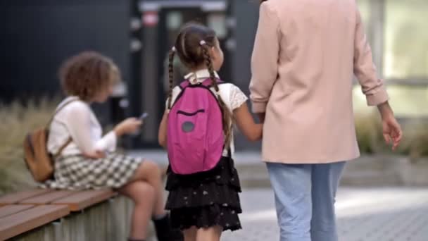 Una mujer adulta lleva a su pequeña hija de la mano a la escuela. Una adolescente con un teléfono inteligente en sus manos está sentada en el banco. Días escolares. — Vídeo de stock