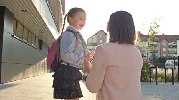 Mujer joven acompaña a su hija pequeña a la escuela. La colegiala besa a su madre adiós y felizmente corre a clase. — Vídeo de stock