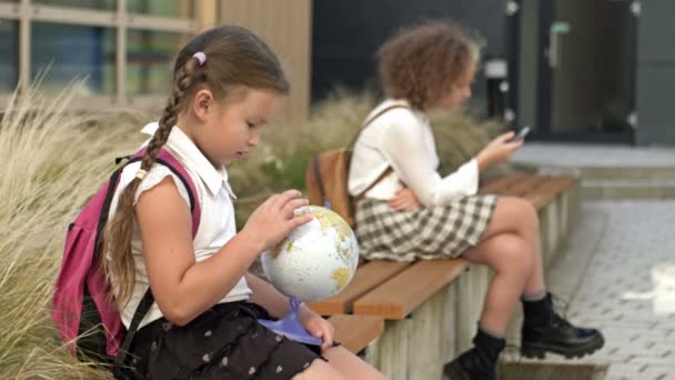 Dvě školačky různého věku sedí na lavičce na školním dvoře. Holčička si hraje s glóbem, nejstarší má v rukou smartphone. — Stock video