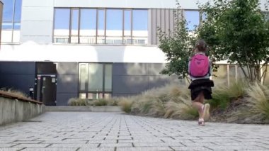 Bir ilkokul öğrencisi aceleyle okula gider. Okul binasının arka planında at kuyruklu ve sırt çantalı küçük bir kız koşuyor..