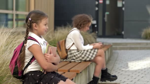 Due studentesse di età diverse sono sedute su una panchina nel cortile della scuola. La bambina preme i libri sul petto, la maggiore ha uno smartphone tra le mani. — Video Stock