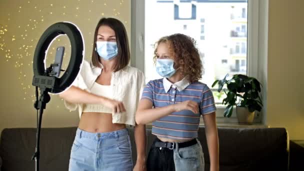 若い母親と保護マスクの大人の子供がカメラの前で踊り、ビデオを撮影しています。covid-19コロナウイルスからの隔離と自己分離の利点をお楽しみください。. — ストック動画