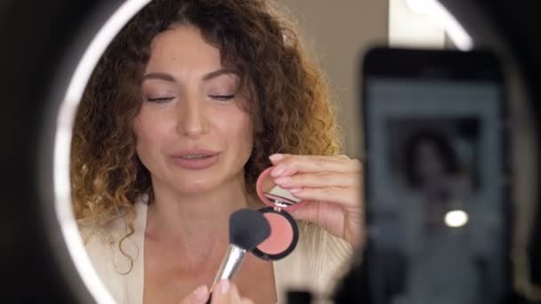 Goed verzorgde, middelbare leeftijd beauty blogger praat over de techniek van het aanbrengen van blush op haar gezicht. Een vrouw zit voor een spiegel en een videocamera. — Stockvideo