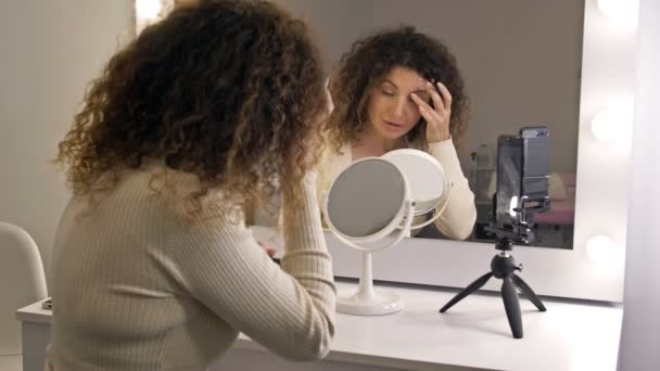 İyi bakımlı, orta yaşlı güzellik blogcusu göz makyajından bahsediyor. Aynanın önünde oturan bir kadın ve bir video kamera.. — Stok video