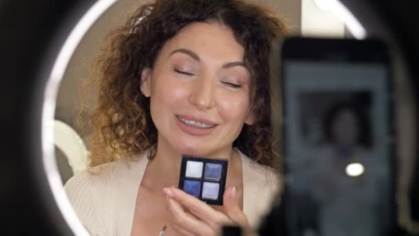 Goed verzorgde, middelbare leeftijd beauty blogger praat over modetrends in oogmake-up. Een vrouw zit voor een spiegel en een videocamera en toont een doos met oogschaduw. — Stockvideo