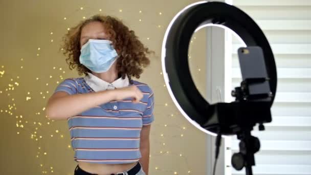 의료용 마스크를 쓴 채 카메라 앞에서 춤을 추는 즐거운 소녀는 소셜 미디어를 통해 자신의 인기있는 콘텐츠를 모바일 앱으로 만들어 냈다.. — 비디오