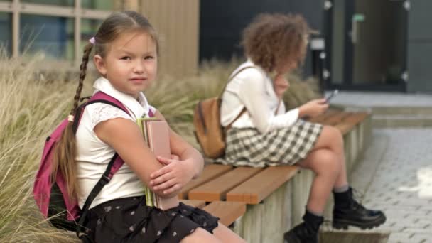異なる年齢の2人の女子学生が学校の庭のベンチに座っています。少女は本を胸に押し当て、長老はスマートフォンを手に. — ストック動画