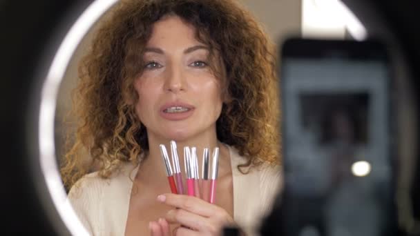 Schöne Beauty-Bloggerin mittleren Alters spricht über Lippenstifte. Frau sitzt vor einem Spiegel, in dem sich eine Videokamera spiegelt. — Stockvideo