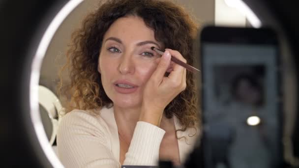 Välvårdad, medelålders skönhetsbloggare pratar om ögonmakeup. En kvinna framför en spegel och en videokamera. — Stockvideo