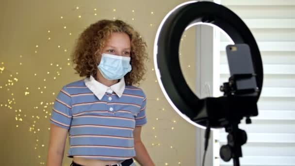 Χαρούμενη έφηβη που χορεύει στην κάμερα με μια ιατρική μάσκα κινηματογραφώντας βίντεο, δημιουργώντας το μοντέρνο περιεχόμενό της σε μια εφαρμογή για κινητά για να το μοιραστεί στα μέσα κοινωνικής δικτύωσης. — Αρχείο Βίντεο