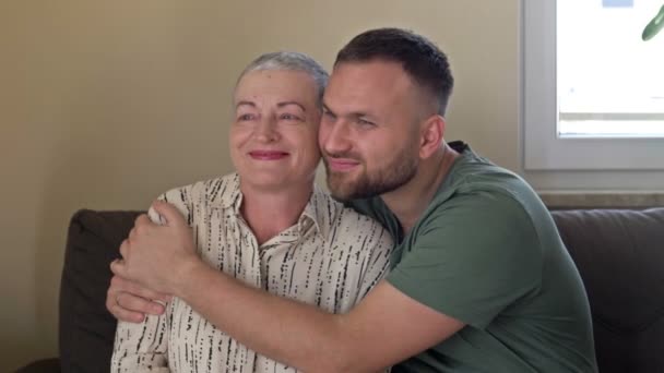 Ein erwachsener Sohn umarmt seine Mutter. Eine ältere Frau hat nach einer Chemotherapie Nachwachsen der Haare. Liebe, Fürsorge und Unterstützung in der Familie eines Krebspatienten. — Stockvideo