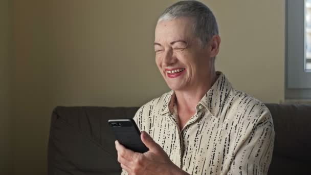 En äldre kvinna tittar på smartphone skärmen och skrattar mycket glatt och uppriktigt. — Stockvideo