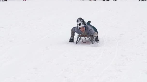Veselý chlapec v lyžařském obleku klouže po svahu na saních, pomáhajících si rukama. Dítě je šťastné.. — Stock video