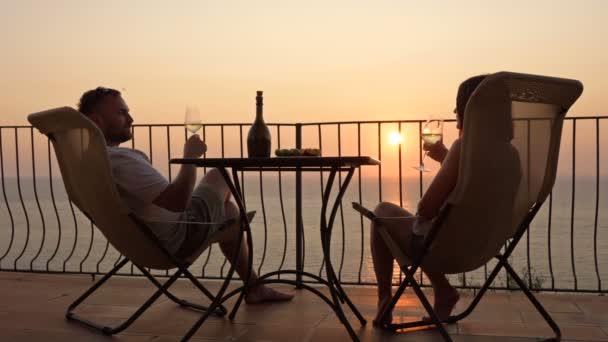 Hermosa pareja joven bebiendo vino en la terraza y admirando la puesta de sol sobre el mar. — Vídeos de Stock