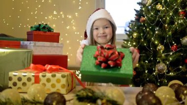 6-7 yaşlarında Noel Baba şapkası takan bir kız Noel ağacının arka planında ve hediyelerle dolu parlak kutular. Çocuk birine içinde Noel hediyesi olan bir kutu verir..