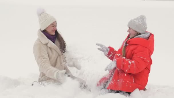 Young woman with her teenage daughter is kneeling in the snow and cheerfully sprinkling snow on each other. Winter fun. — Stock Video