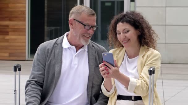 Mooi volwassen stel met koffers zit op een bankje te wachten op hun vlucht. Een man en een vrouw bespreken wat ze zagen op het scherm van een mobiele telefoon.. — Stockvideo