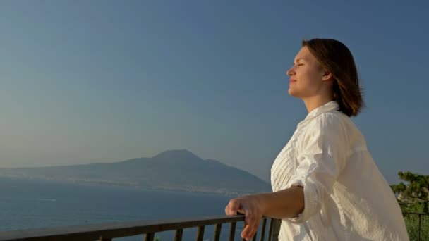 Молодая женщина стоит на балконе с прекрасным видом на море и горы. Женщина с радостью обнажает свое лицо под лучами солнца. — стоковое видео