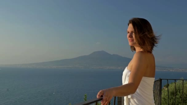 Jeune femme se dresse sur un balcon avec une belle vue sur la mer et les montagnes. Femme expose volontiers son visage aux rayons du soleil. — Video