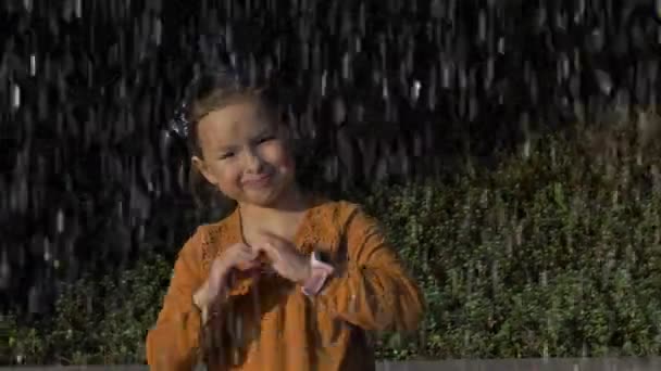 Menina 6-7 anos atrás de uma cortina de spray de fonte. Criança envergonhada mostra o coração com um gesto. — Vídeo de Stock