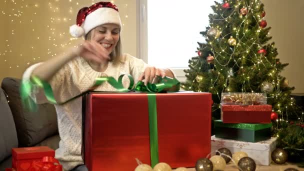 Donna affascinante in cappello Babbo Natale avvolge amorevolmente regali per i propri cari. Sullo sfondo di un albero di Natale. Buon Natale. Ciao.. — Video Stock