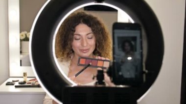 İyi bakımlı, orta yaşlı güzellik blogcusu göz makyajındaki moda eğilimlerinden bahsediyor. Bir kadın aynanın ve kameranın önünde oturur ve göz farı olan bir kutu gösterir..