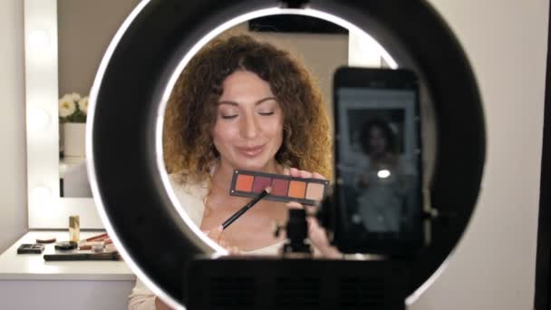 El bloguero de belleza bien cuidado y de mediana edad habla sobre las tendencias de la moda en el maquillaje de ojos. Una mujer se sienta frente a un espejo y una cámara de video y muestra una caja con sombra de ojos. — Vídeo de stock